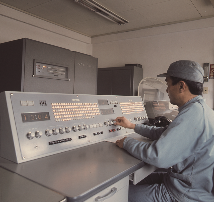 Man in scrub coat with hard hat sitting at an older computer panel.