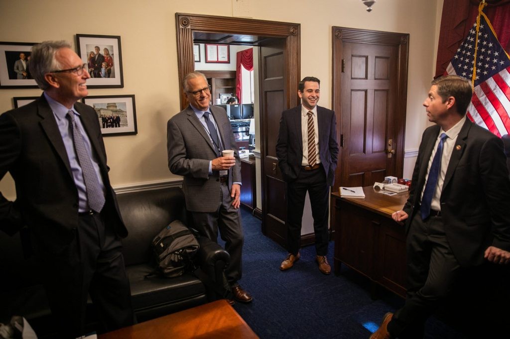 four men talking and standing in congressional office with American flag in background