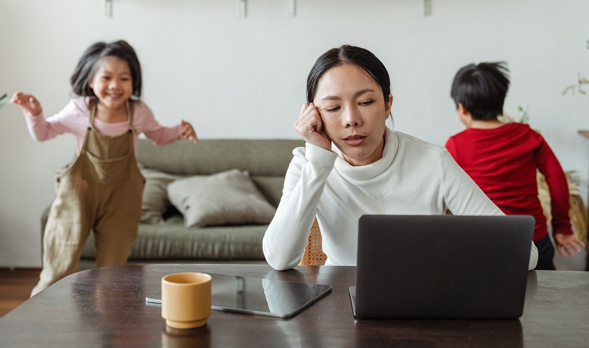 overwhelmed mom on laptop while two kids are running around the room