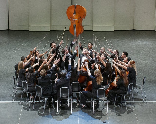 group photo of one of McQueen High School's orchestra all holding bows up as director Kenny Baker raises a cello in the air