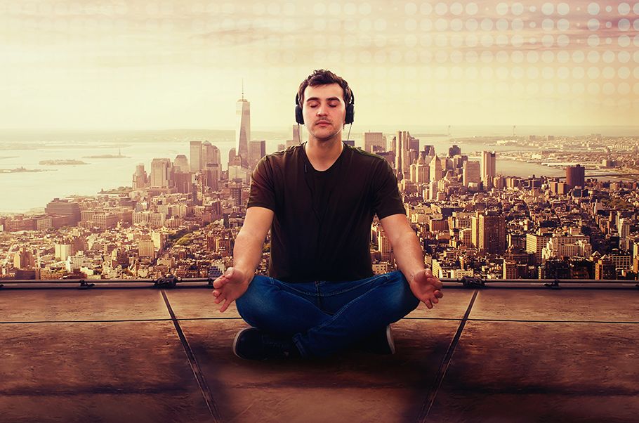 Man sitting in a lotus position meditating on on a roof with a cityscape behind him. He is wearing headphones.