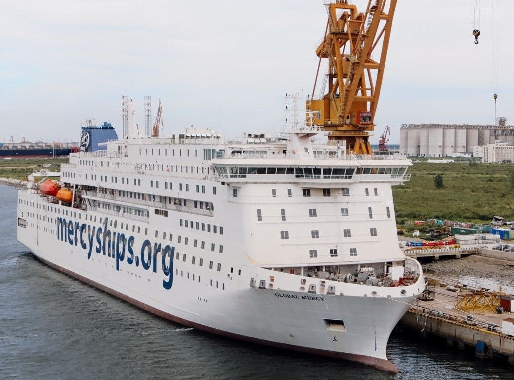 Large white passenger ship in dock with a crane in the background. The words "mercyships.org" is painted on the side of the ship.