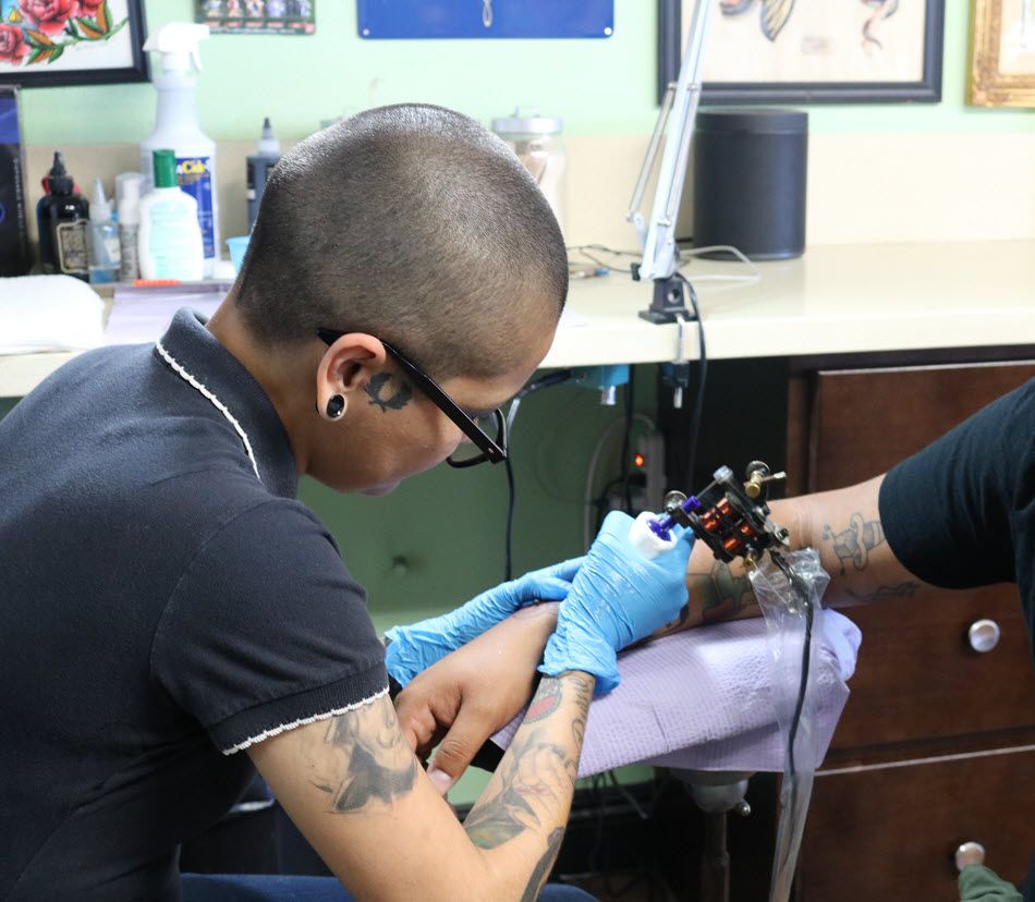 Closeup of tattoo artist inking someone's arm. The speaker from adjacent closeup photo is visible on counter in background.