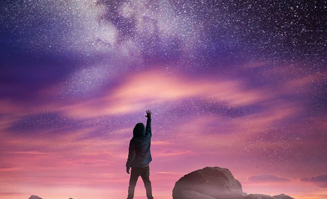 Person standing with back to camera in silhouette on top of a rocky hill with one hand reaching out towards the starry sky.