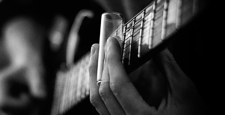 Closeup black and white image of someone playing guitar with a slide. In foreground is the hand on the guitar neck and on the middle finger is a glass slide.