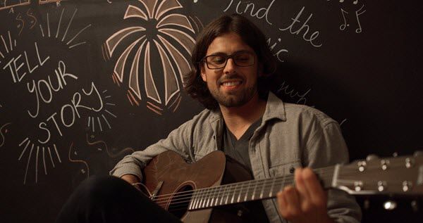 Man who appears to be in his 30s dressed in casual clothes smiling while playing an acoustic guitar. His back it to a blackboard where there are different sayings written including "Tell your story".