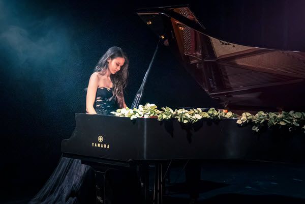 Young woman in ball gown seen in profile playing a Yamaha concert grand piano on darkened stage.
