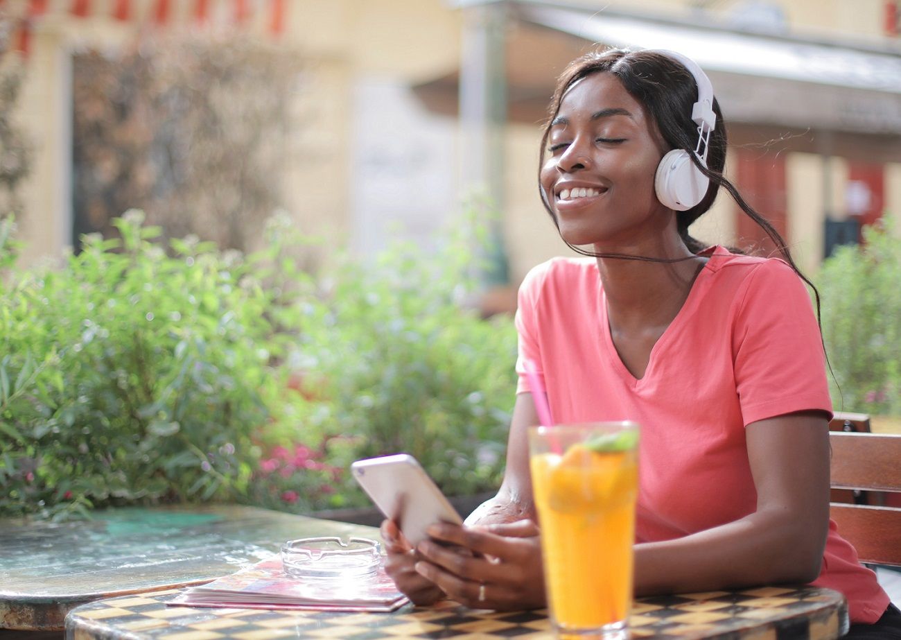 Black woman, smiling with eyes closed and listening to music sitting at an outdoor table with a glass of juice