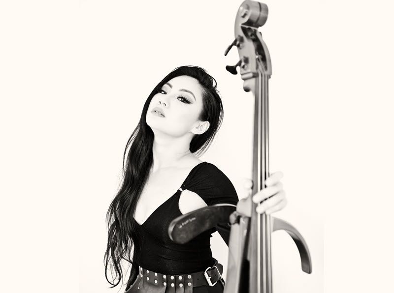 Young Asian woman with long hair, black shirt and leather skirt holding the neck of an open bodied electric cello.
