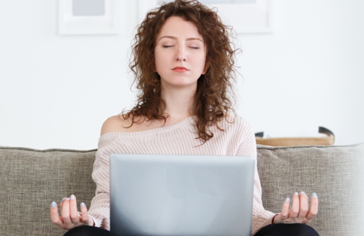 Woman sitting crosslegged on a couch meditating with a laptop open in her lap.