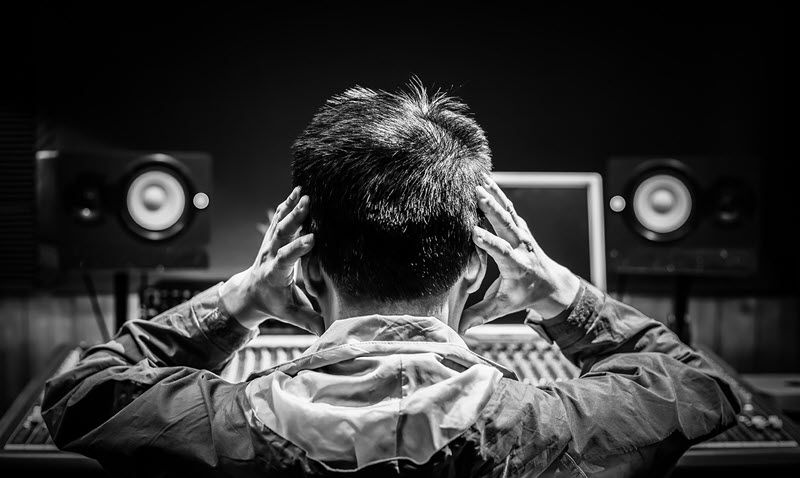 View from behind of a man putting his hands where his headset would be and is seated in front of a mixing board.