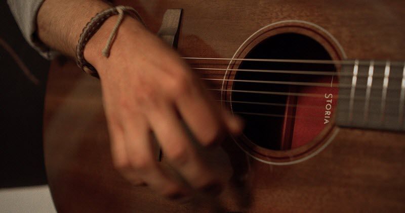 Closeup of a hand playing an acoustic guitar. Through the guitar hole, there is a label with the name "Storia".