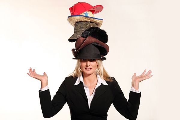 Woman in business attire with many hats stacked on top of her head.