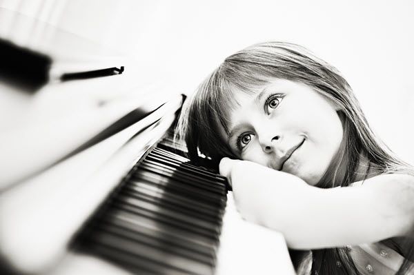 Black and white image of a young girl smiling while resting her arm and head on her piano keys.