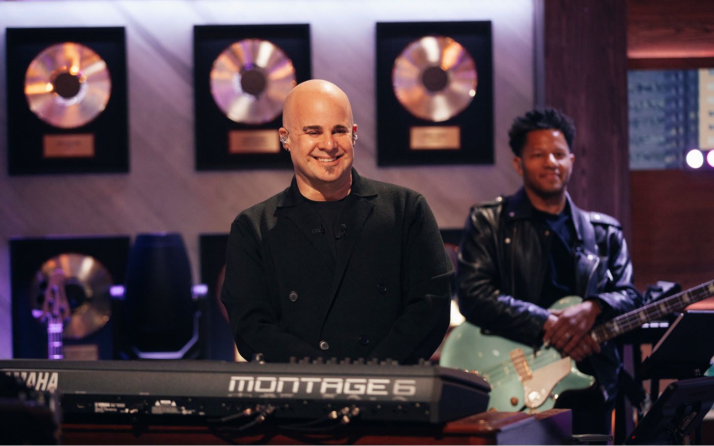Attractive bald male musician with a guitarist in the background an a wall of framed gold albums smiles for camera.