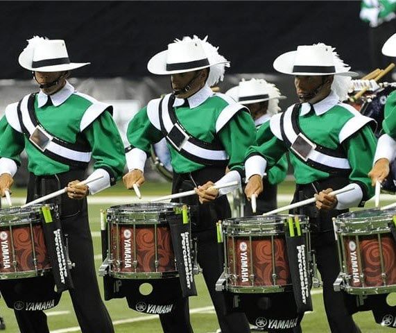 A collegiate drumline marching on a field in uniform each playing a Yamaha snare drum.