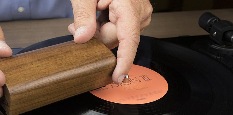 Closeup of man's hands as he uses a small rectangular object to clean a vinyl record on a turntable.