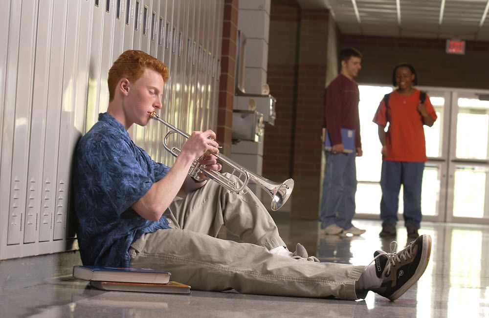 Teenage boy sits on school floor with back against lockers playing his trumpet while two other students down the hall listen