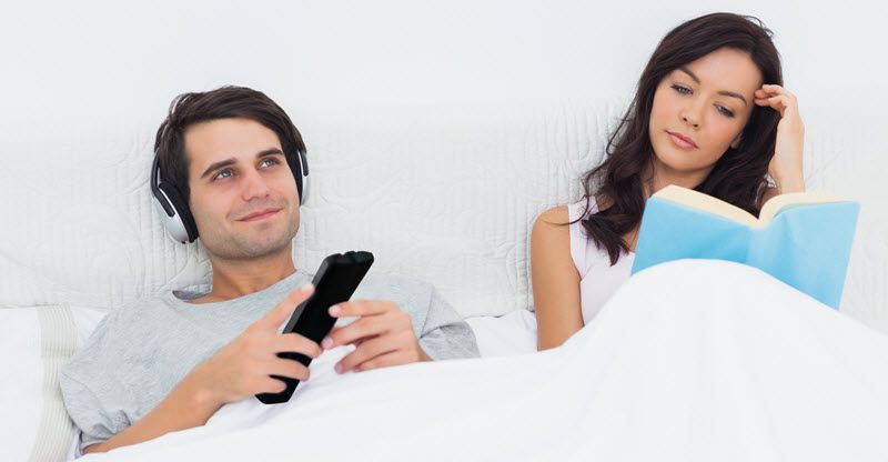 Woman reading in bed while her husband is wearing headphones with TV remote in his hand.