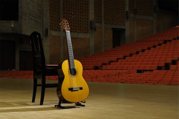 Classical guitar upright on a stand next to an empty wooden chair center stage in a large empty concert hall.