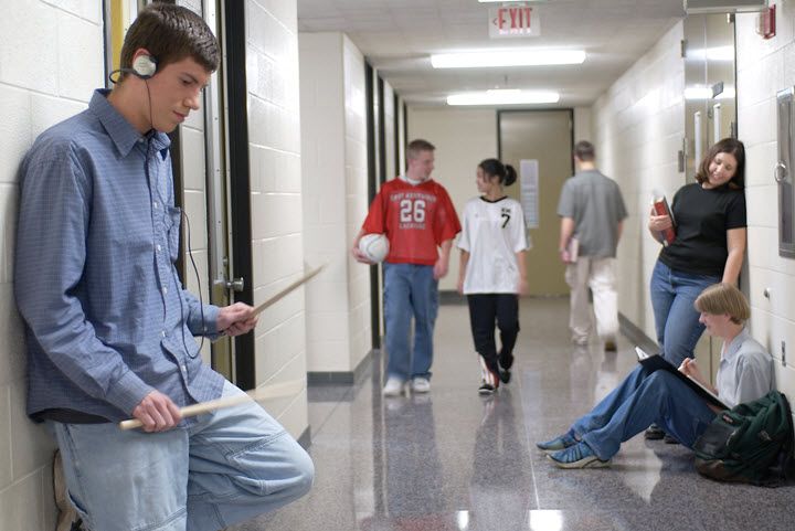 Young man leaning against a school hallway wall wearing headphones and drumming with snare drumsticks in the air while other students chat and stroll in the hallway undisturbed.