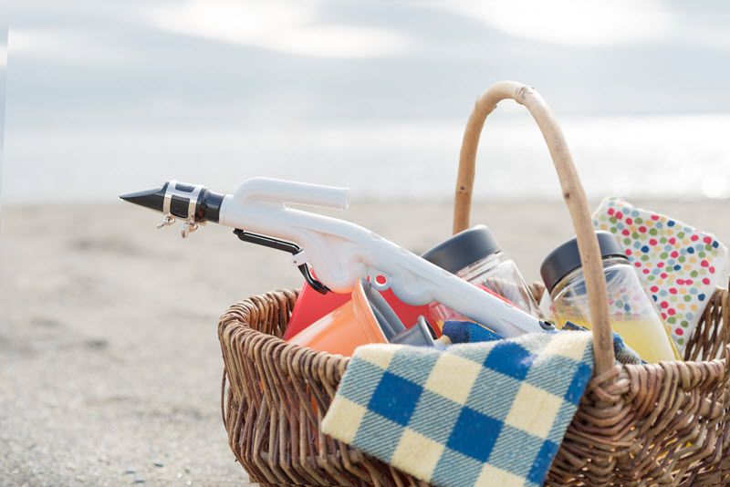 Picnic basket on sand on the beach with various jars and tablecloth and a plastic reeded instrument sticking out of the top of the basket.