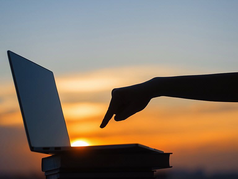 Hand poised with finger extended above a laptop keyboard ready to press a key with the sun going down in background.