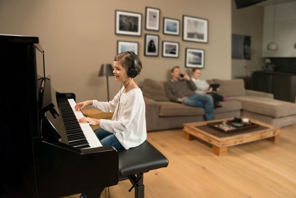 Young girl practicing the piano in the living room with a headset on while her parents are relaxing in background talking on mobile and working on a tablet.