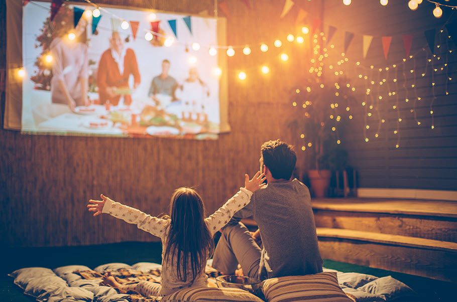 A child sits on a blanket in the backyard next to dad watching a moving projected on a sheet on a wall.