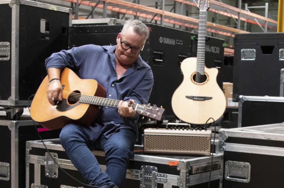 Man playing an acoustic guitar while sitting backstage with small amp adjacent.