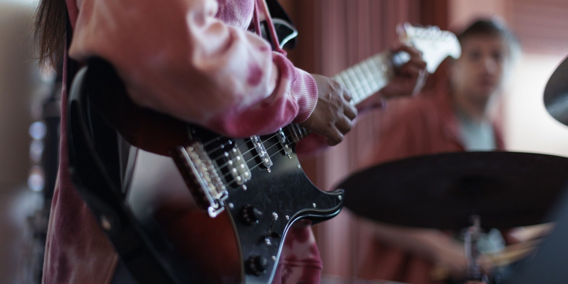 closeup of someone playing an electric guitar with a drum set player in the background