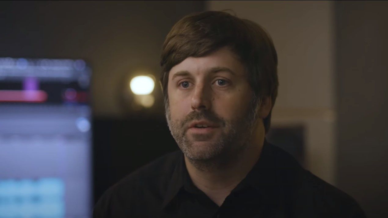 Man in his 30s with short hair, moustache and beard looking at camera. There is a digital studio setup behind him.