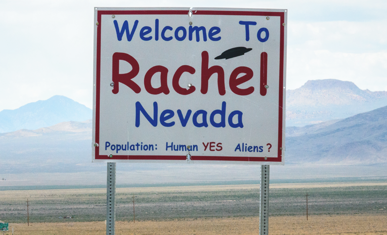 A photo of the "Welcome to Rachel Nevada" road sign with the long road leading into the desert and mountains beyond that to the left of sign. On sign, it indicates that "Population: Human YES and Alien ?".
