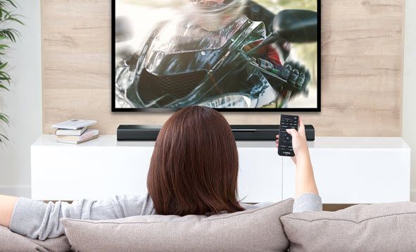 Woman seen from behind sitting on a couch in modern living room with remote facing a flat screen with sound bar below TV.