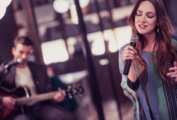 A young woman on stage singing into a microphone with a young man playing guitar in the background.