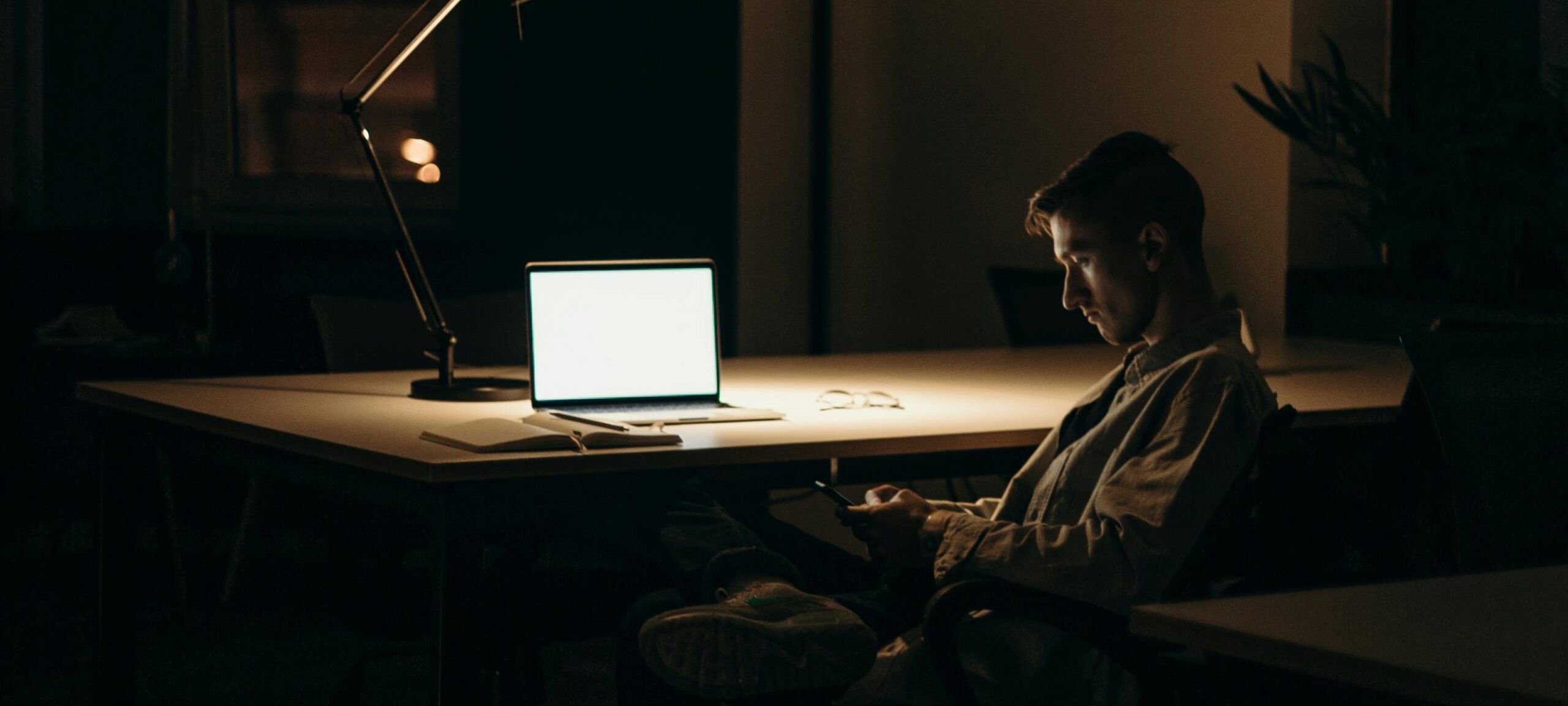 man looking at phone while sitting at desk with an open laptop