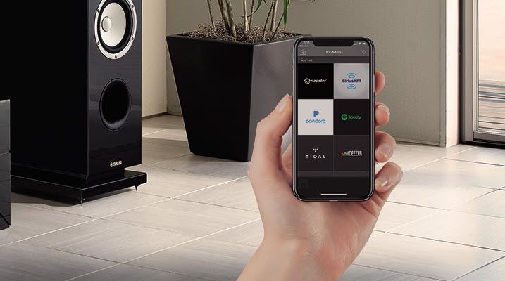 Living room scene with speakers and sound system in background and someone's hand in foreground holding a smartphone with app showing.