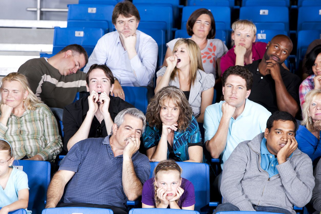 Audience in stadium seats with expressions of boredom on faces and in bodies: resting heads on hands, sleeping or yawning.