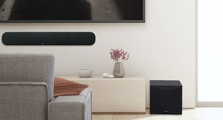 Modern living room with subwoofer on floor and a sound bar installed on wall below flat screen facing couch.