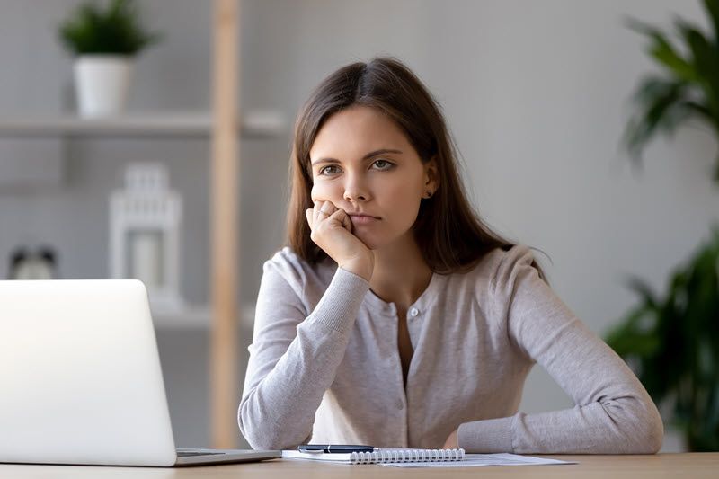 Woman sitting at her home computer desk with her chin resting on her hand and just staring at the computer screen.
