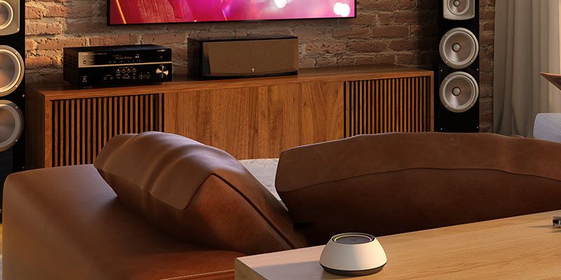 Living room with a couch facing an audio system. Behind the couch closest to the camera is a table with a cylindrical smart home element visible.