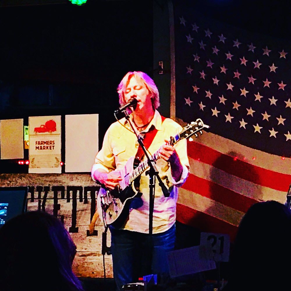 Man singing and playing guitar at a microphone on a small stage. There is an American flag hanging on wall behind him.