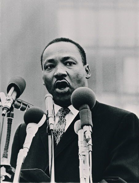 Dr. Martin Luther King speaking behind four news microphones.