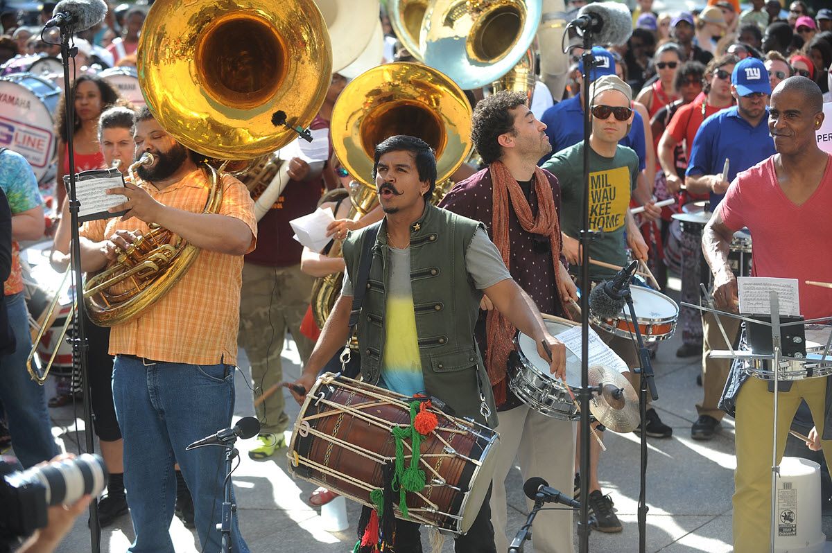 Percussion and horn band playing on a sidewalk with a large crowd enjoying the show.