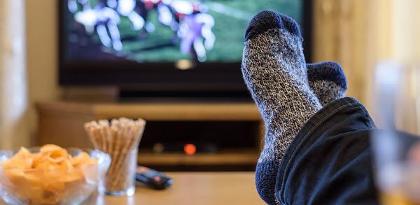 Someone's feet on a coffee table with chips and pretzels with a football game on a large flat screen in background.