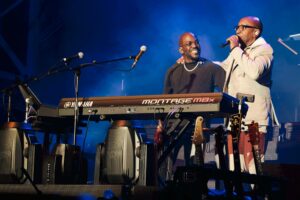 A man in a white suit onstage talking into a microphone with his arm around another man playing synthesizer keyboard.