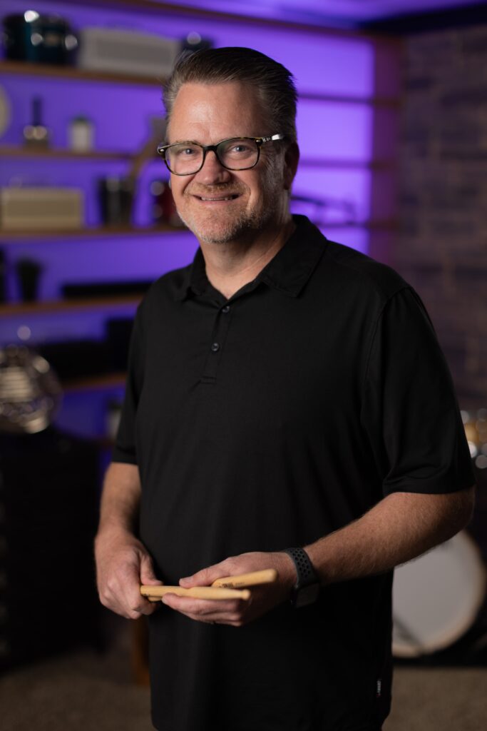 A smiling man holding a pair of drumsticks.