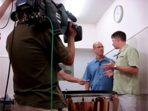 Two men being filmed talking as they stand over a timpani.