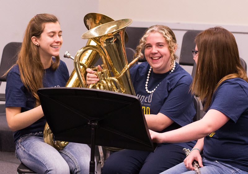 Three teenage girls in band class laughing while sharing music stand and holding instruments.