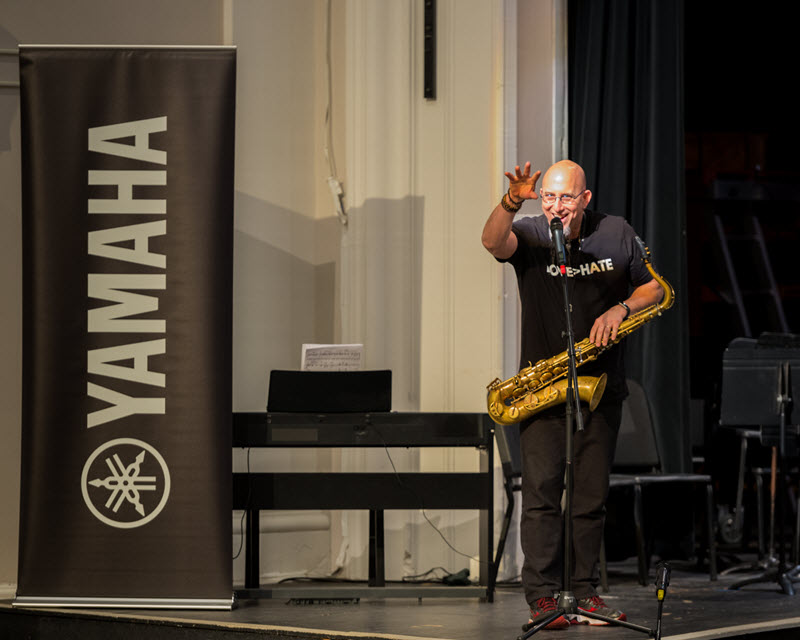 Saxophonist Jeff Coffin holding his saxophone while speaking at a microphone on a school stage with the banner on the left showing Yamaha logo.